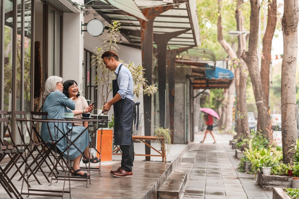 Two senior women are using their phone to order and pay for their coffee at a cafe. The waiter is using a digital tablet.