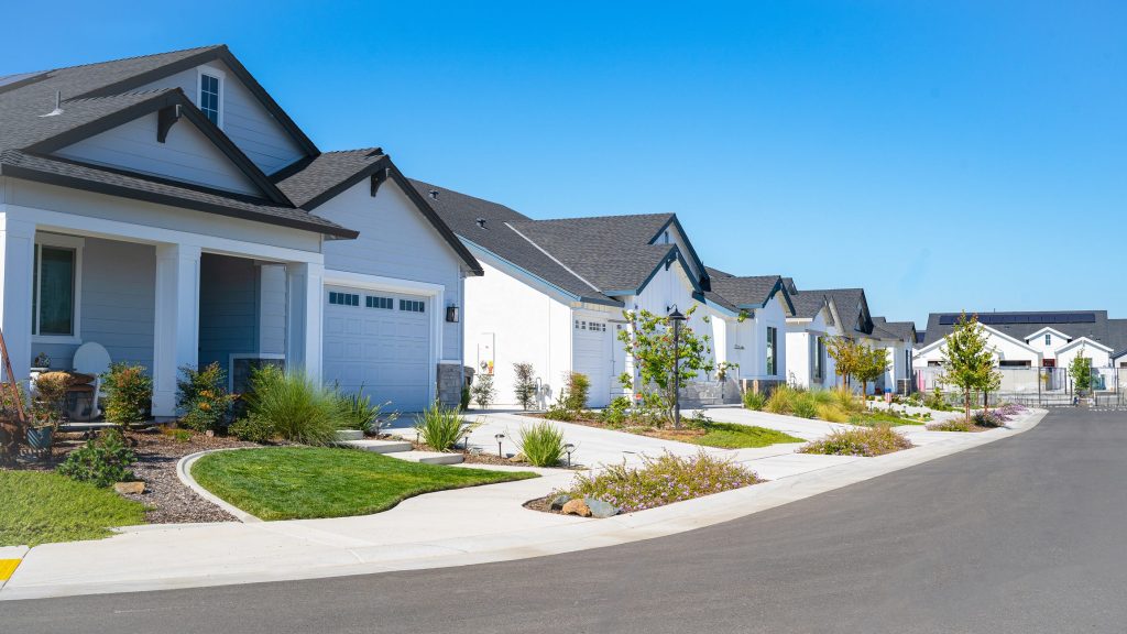 A suburban residential street with modern, white houses on both sides, manicured lawns, and a clear, blue sky.