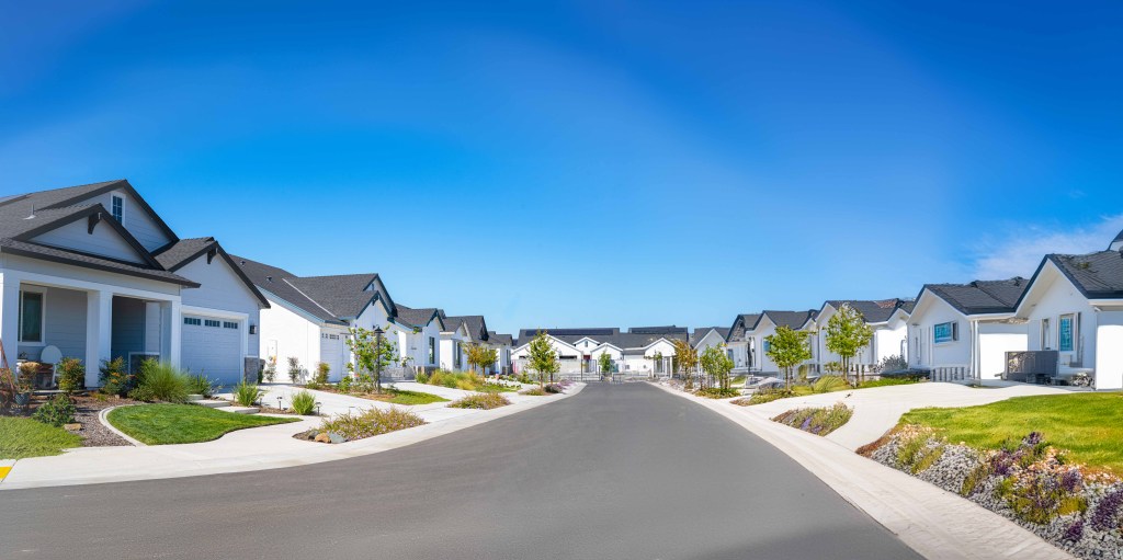 A suburban residential street with modern, white houses on both sides, manicured lawns, and a clear, blue sky.
