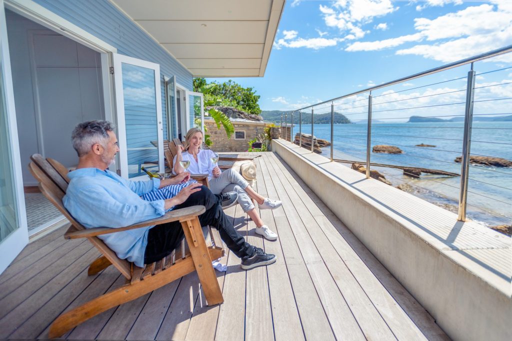 Mature couple drinking wine out on the deck in their waterfront home. They are happy and smiling sitting in deck chairs. The sea is in the background