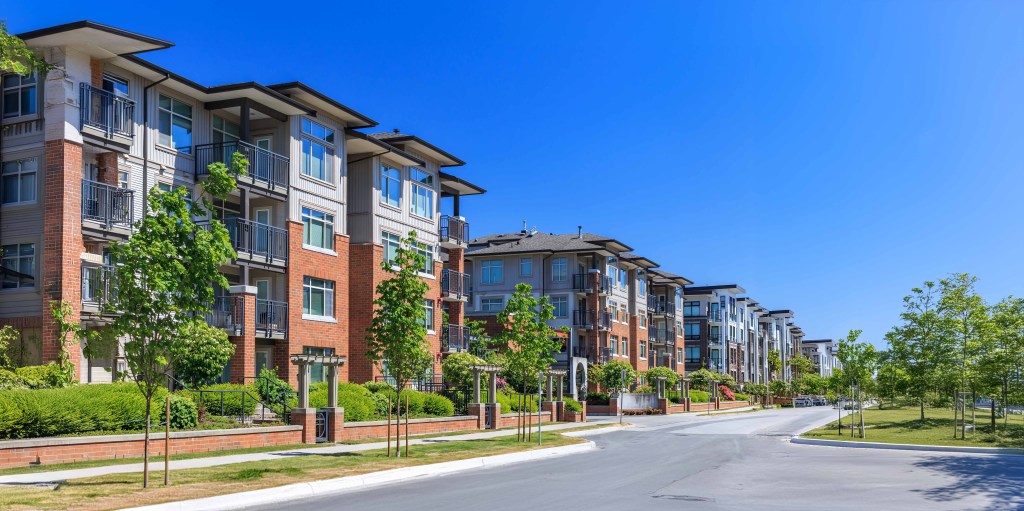A sunny day with a clear blue sky over a residential street lined with modern apartment buildings featuring multiple floors and balconies. Young trees and well-manicured landscaping are seen along the sidewalk.