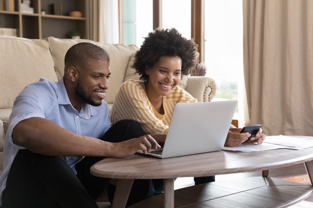 Happy young couple using laptop, looking at screen, smiling, laughing, getting good news. Husband and wife using online banking app on computer, paying bills, insurance on the internet