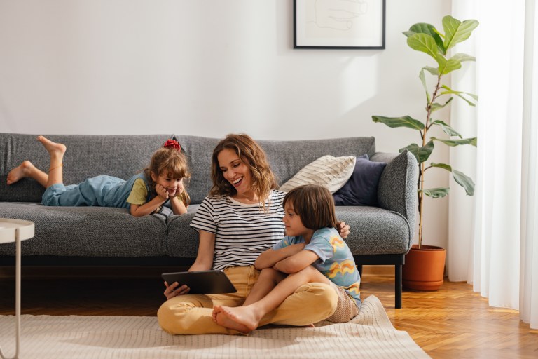 Mother with her kids sitting on the floor and using digital tablet for playing games, watching movies and relaxing at home. Her son is cuddled with her, while the daughter is lying on the sofa next to them.